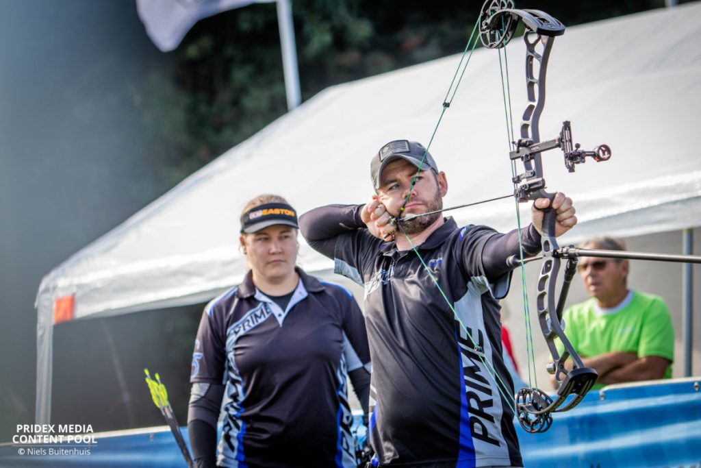 Een man richt een compoundboog terwijl een vrouw achter hem staat, beiden in sportkleding, tijdens een handboogschietevenement in de open lucht met een tent op de achtergrond.