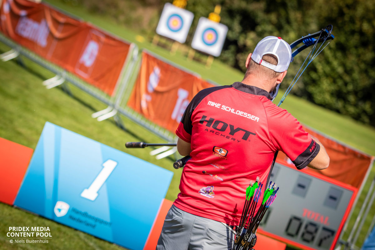 Een boogschutter bereidt zich voor om een pijl naar doelen te schieten tijdens een handboogschietwedstrijd, gekleed in een rood Hoyt Archery-shirt. Op een scorebord staat het nummer 087 en aan de linkerkant is een groot '1'-teken zichtbaar.