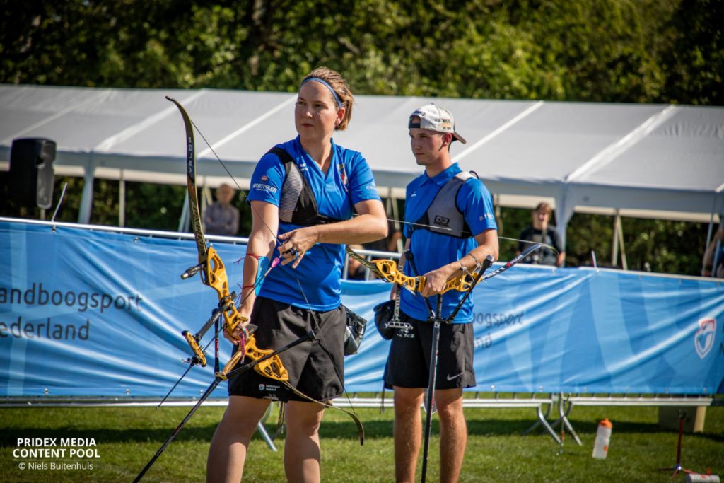 Twee boogschutters, een vrouw en een man, in blauwe uniformen met bogen, staan op een grasveld tijdens een handboogschietwedstrijd. Witte tenten en een blauwe banner zijn op de achtergrond.