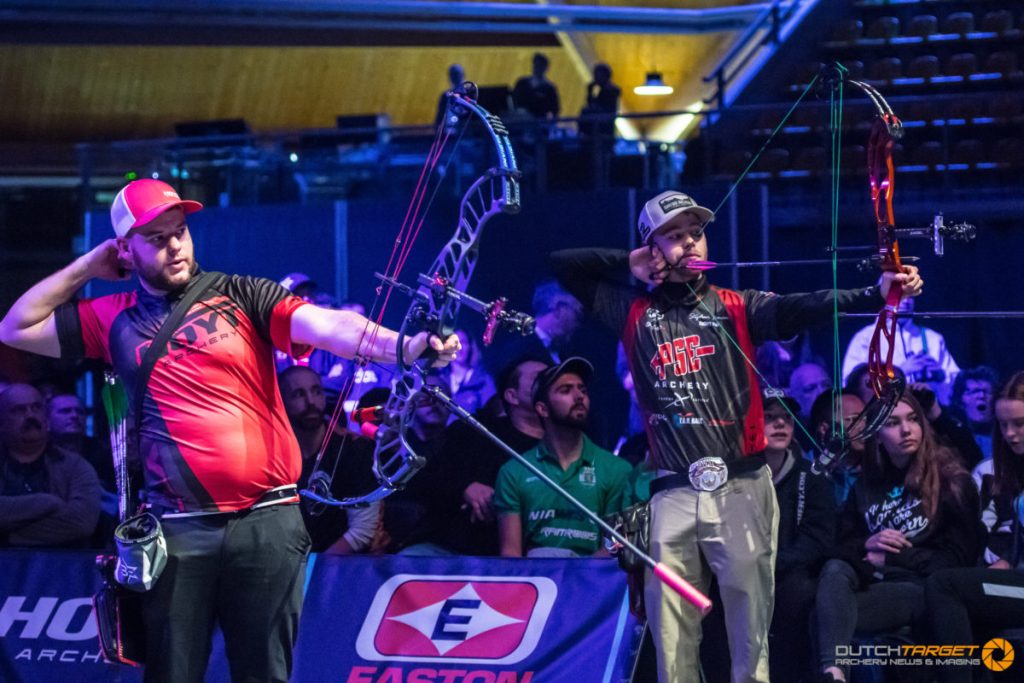 Twee boogschutters, die binnenshuis naast elkaar staan, richten hun compoundbogen tijdens een competitief handboogsportevenement. Het publiek kijkt vanaf de achtergrond toe.