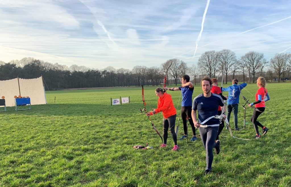 Mensen die deelnemen aan een handboogsportevenement op een grasveld. Sommigen bereiden zich voor om hun pijl af te schieten, terwijl anderen weglopen van de doelen. De lucht is helder met zichtbare dampsporen.