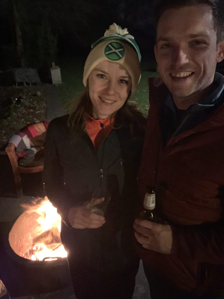 Twee mensen zijn 's nachts buiten naast een vuurplaats. De persoon links draagt een hoed en houdt een drankje in de hand, terwijl de persoon rechts, net na een middagje handboogschieten, een fles vasthoudt.