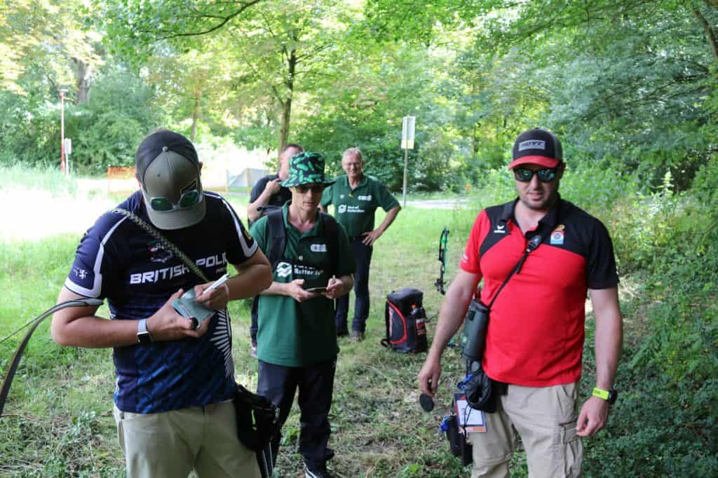 Buiten staat een groep van vijf boogschutters. Twee staan op de voorgrond, gekleed in sportkleding en hoeden, gefocust op hun pijl-en-booguitrusting, terwijl drie anderen op de achtergrond bij een bosrijke omgeving staan.