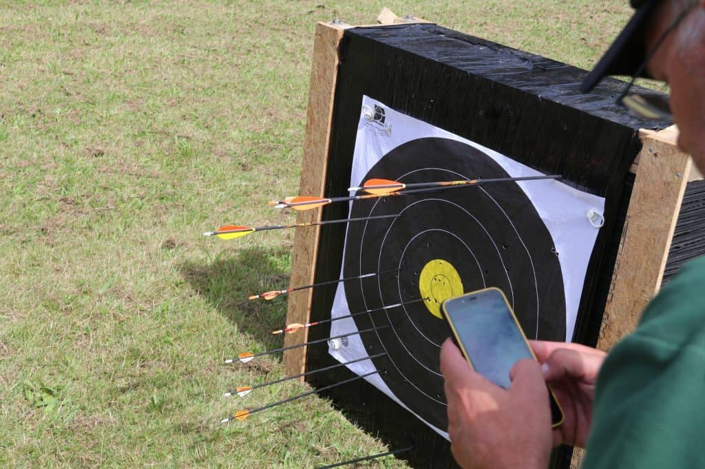Een persoon gebruikt een smartphone om boogschietscores vast te leggen naast een doelwit met pijlen – waarvan er meerdere het gemarkeerde gebied raken – op een grasveld, ondergedompeld in de opwinding van handboogschieten.