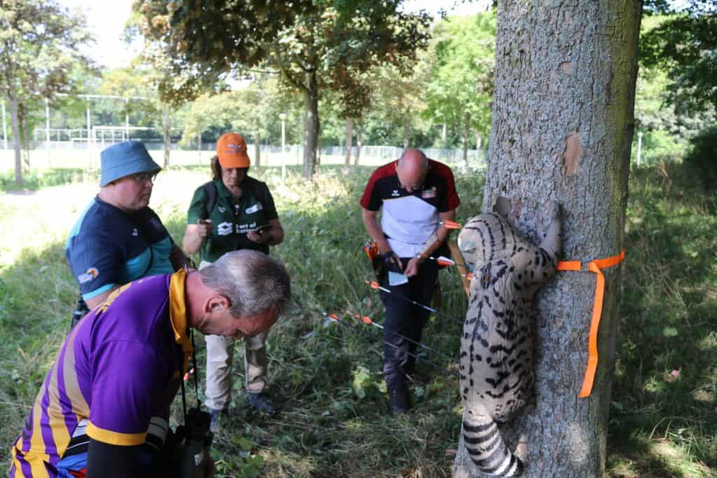 Verschillende mensen verzamelen zich in een buitenomgeving, observeren een boom met een levensecht dierendoel eraan en oefenen handboogschieten.