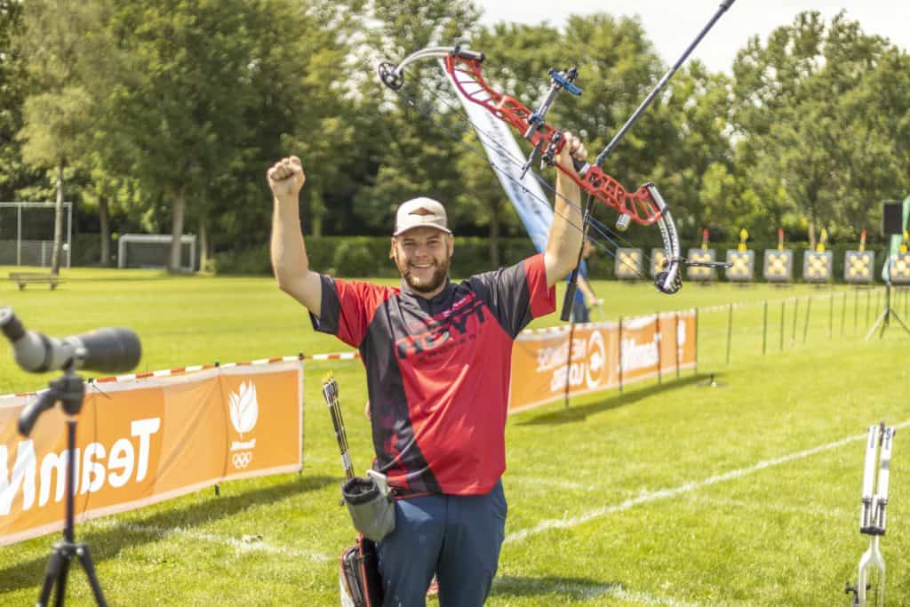 Een boogschutter in een rood-zwarte outfit heft feestelijk zijn arm op een zonnig veld met schietborden en spandoeken op de achtergrond, waarmee hij zijn handboogschietvaardigheid laat zien.