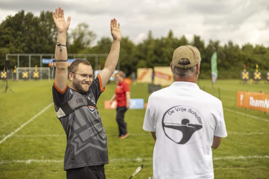 Man in grijze sporttrui heft zijn armen feestelijk op terwijl een andere man in een wit overhemd met "De Vrije Schutter" op de rug toekijkt. Op de achtergrond is een buitenboogschietveld zichtbaar, gewijd aan de handboogsport.