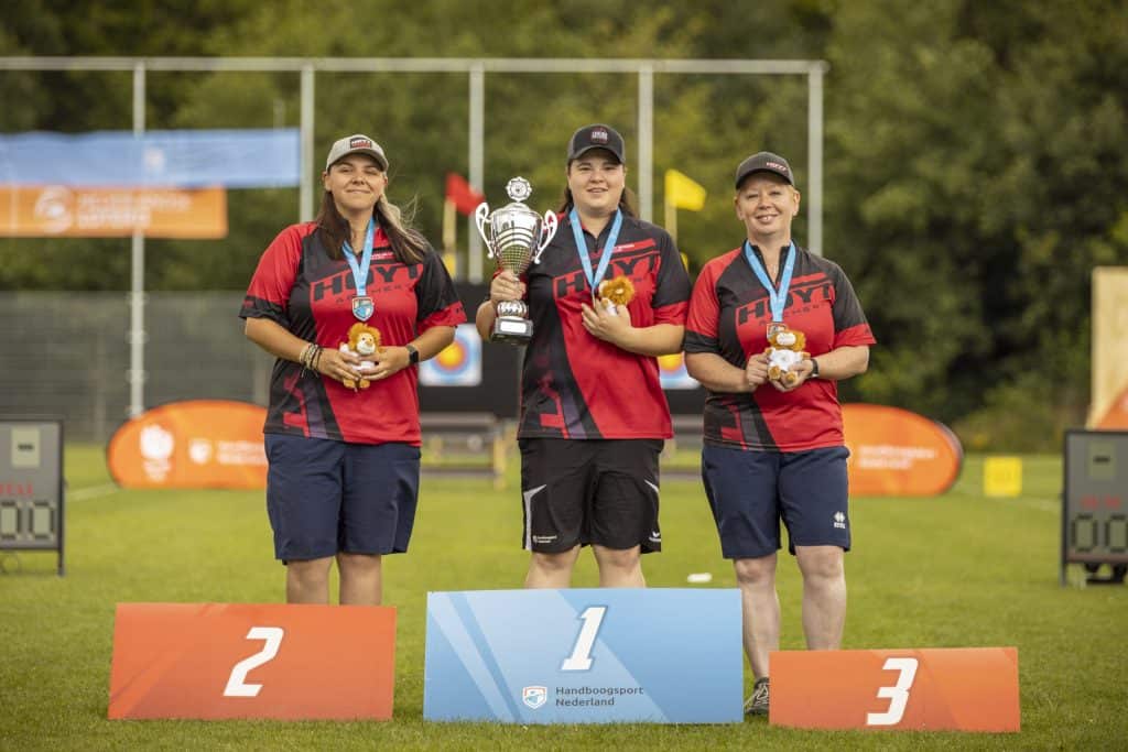 Drie boogschutters, staande op podia met de nummers 2, 1 en 3, houden medailles en trofeeën vast van een handboogschietevenement. Ze dragen allemaal bijpassende outfits en de winnaar van de eerste plaats heeft een grotere trofee.