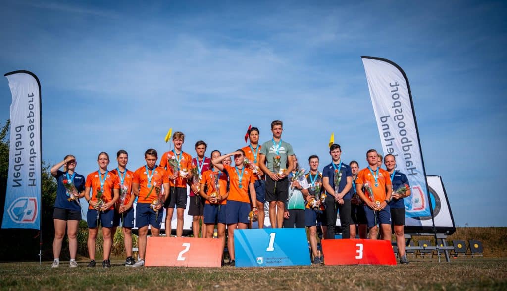 Atleten staan op podia met medailles en trofeeën tijdens een handboogsportevenement in de open lucht, drie op het bovenste podium en zes op elk van het tweede en derde podium. Op de achtergrond zijn vlaggen en spandoeken te zien.