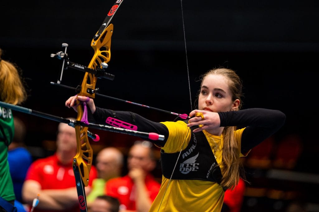 Een jonge vrouwelijke boogschutter in een gele top richt geconcentreerd haar pijl tijdens een indoor handboogschietwedstrijd, met wazige toeschouwers op de achtergrond.