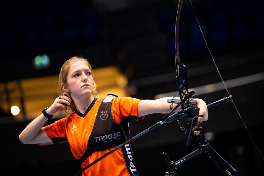 Een persoon in een oranje shirt trekt een boogpees naar achteren en richt een pijl tijdens een boogschietevenement. De achtergrond is wazig, waardoor de aandacht wordt gevestigd op de handboogschietende schutter.
