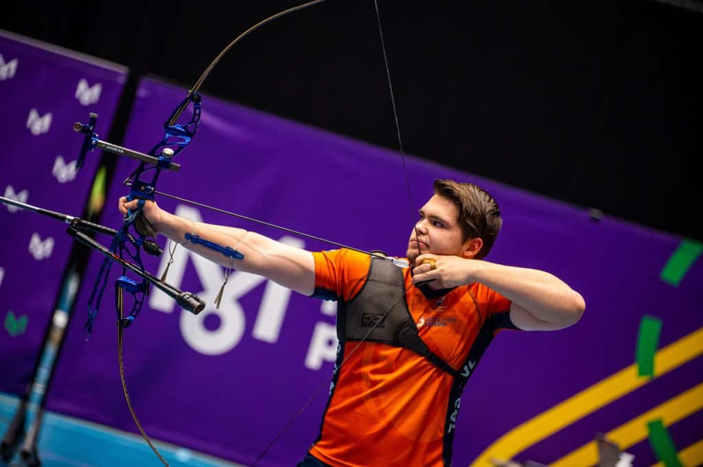 Een boogschutter in oranje shirt trekt geconcentreerd zijn pijl, gericht op een doel. Op de achtergrond is een paarse banner met witte tekst en logo's zichtbaar, die de spanning van het handboogschieten benadrukt.