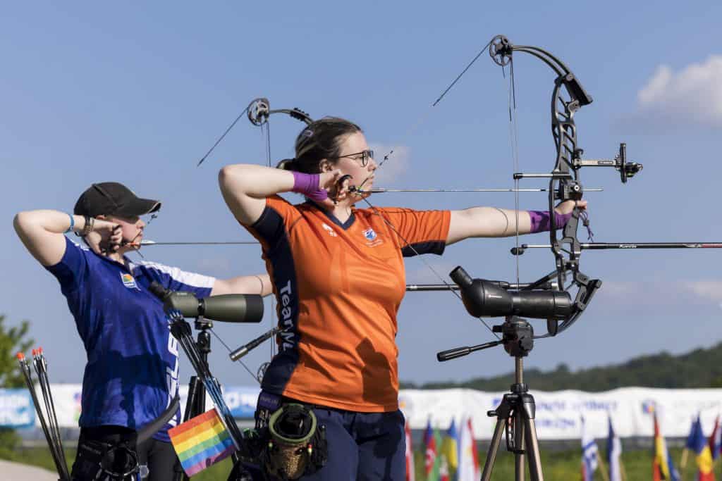 Twee boogschutters in competitieve houding richten hun samengestelde bogen op doelen. De een draagt een blauw shirt en de ander een oranje shirt met het merk "Team NL" op de zijkant. Aan de uitrusting is een regenboogvlag bevestigd, die de inclusieve geest van handboogschieten benadrukt.