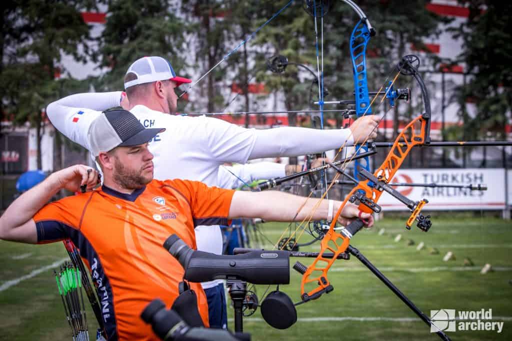 Twee boogschutters richten hun compoundbogen op een grasveld tijdens een handboogsportwedstrijd. De een draagt een oranje overhemd en de ander, in een wit overhemd, trekt de boogpees van zijn boog naar achteren. Op de achtergrond zijn boogschietdoelen te zien.