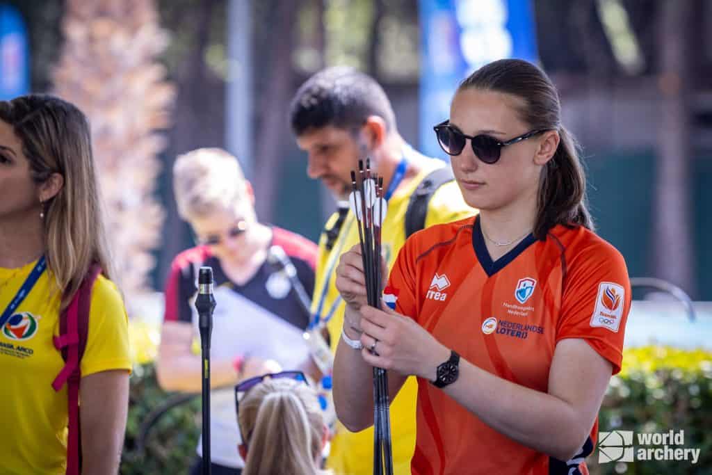 Een boogschutter in een rood shirt organiseert haar pijl terwijl anderen vlakbij staan. De setting blijkt een zonnig handboogsportevenement te zijn. Het World Archery-logo is zichtbaar in de hoek.
