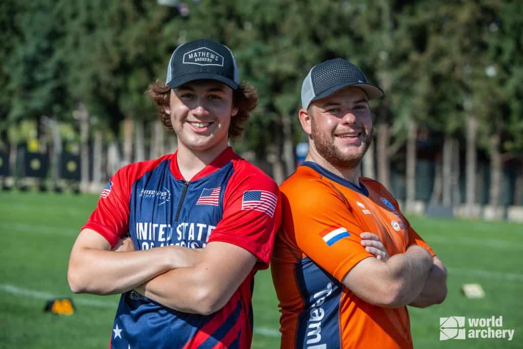 Twee boogschutters staan glimlachend rug aan rug, gekleed in sportuniformen; de ene is rood en blauw met de Amerikaanse vlag, de andere is oranje met een vlag op de mouw. Ieder houdt een boog en een pijl vast, klaar voor handboogschieten. Een groen veld en bomen zijn op de achtergrond.