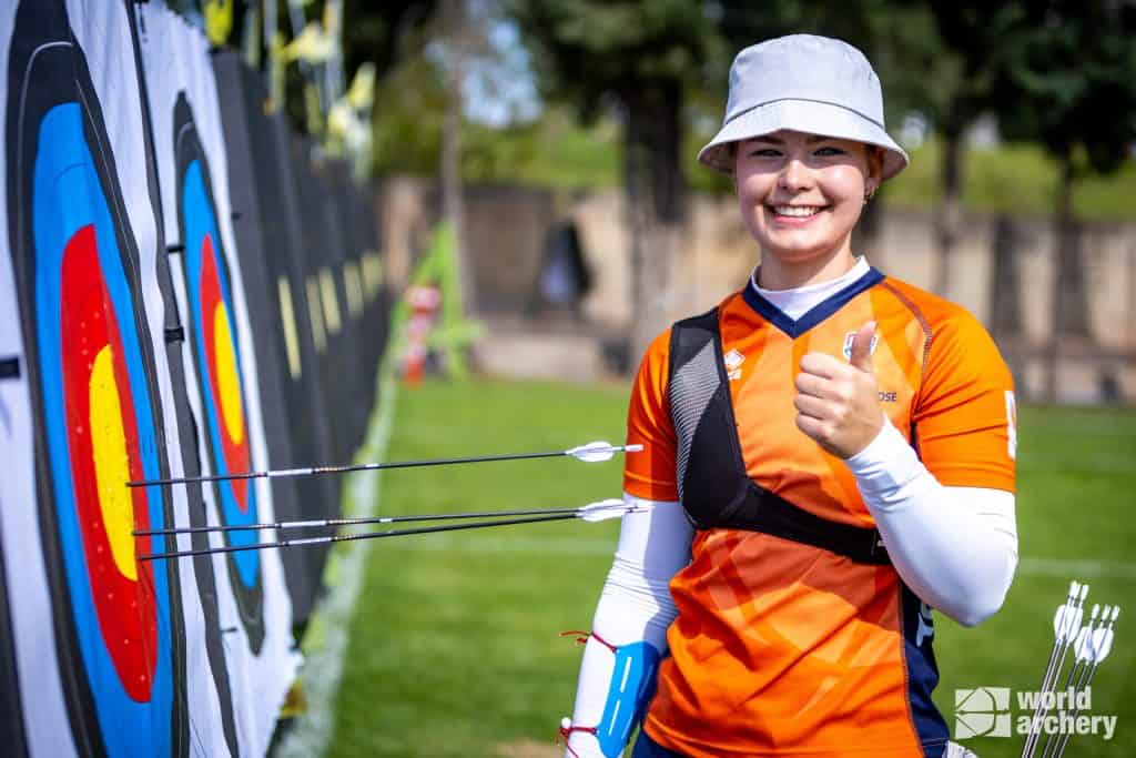 Een boogschutter, gekleed in een oranje en witte outfit en een hoed, steekt zijn duim omhoog terwijl hij naast een doelwit met pijlen staat. Het World Archery-logo is zichtbaar in de rechter benedenhoek en viert de geest van handboogsport.