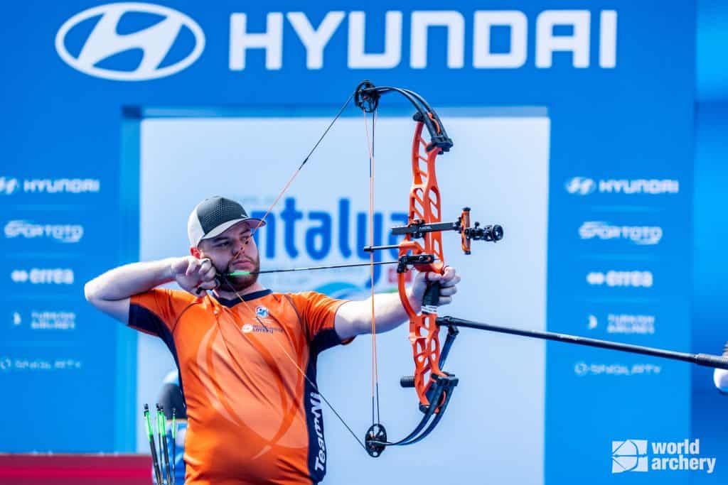 Een man, gekleed in een oranje shirt en hoed, richt een compoundboog tijdens een boogschietevenement, met het Hyundai-logo op de achtergrond. Zijn precieze focus op de pijl benadrukt zijn vaardigheid in handboogschieten.