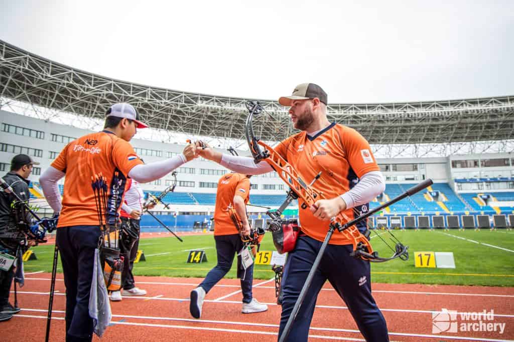 Twee boogschutters in oranje en blauwe uniformen botsen met hun vuist op een baan, terwijl één een pijl en boog vasthoudt. Een andere boogschutter is op de achtergrond. De locatie heeft een groot stadiondak en blauwe zitplaatsen, waardoor de essentie van handboogsport wordt vastgelegd.