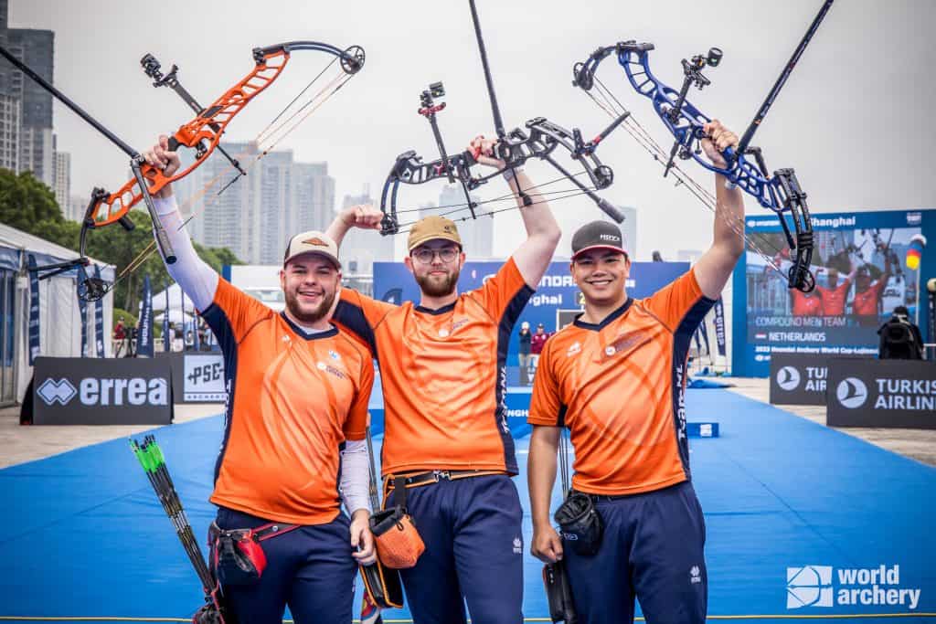 Drie boogschutters in oranje shirts houden glimlachend hun compoundbogen omhoog. Twee van hen hebben medailles om hun nek, die opmerkelijke prestaties in de handboogsport laten zien. Rechtsonder is het logo "World Archery" zichtbaar.