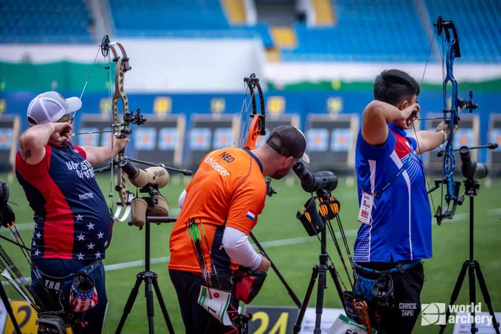 Drie boogschutters in verschillend gekleurde uniformen trekken hun bogen terug en richten hun pijl op doelen tijdens een handboogschietwedstrijd in een stadion.