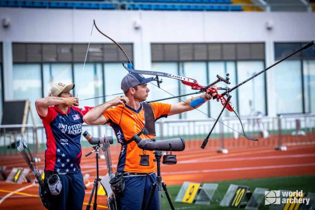 Twee boogschutters richten hun bogen op een buitenbaan, één in een Team USA-trui en de andere in oranje. Aan het einde van het bereik worden doelen gesteld. Ertussen is een spotting scope geplaatst, die de essentie van de handboogsport vastlegt.