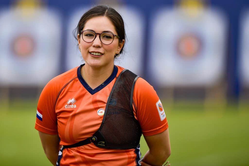 Een persoon met een bril, gekleed in een oranje sportshirt en een borstbeschermer, staat buiten en belichaamt de geest van handboogsport, met vage boogschietdoelen op de achtergrond.
