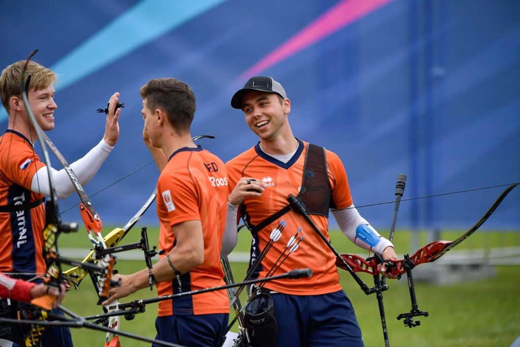 Een groep boogschutters in oranje uniformen, sommigen met pijl en boog in de hand, glimlachend en high-fives gevend in een buitenomgeving.