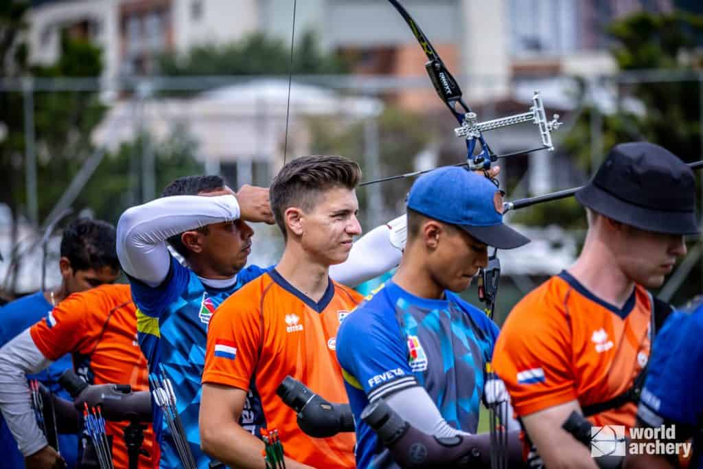 Een groep boogschutters in felgekleurde uniformen staat in de rij en richt hun pijl en boog (pijl). De achtergrond toont een wazig stedelijk landschap. Rechtsonder is het World Archery-logo zichtbaar, dat de essentie van handboogsport weergeeft.
