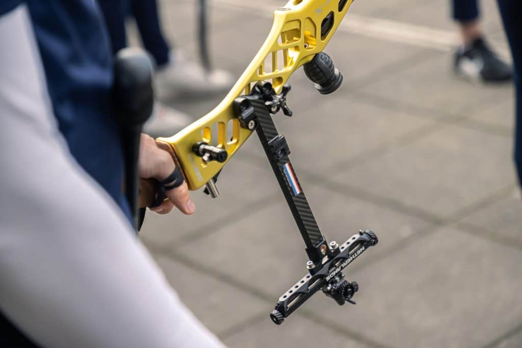 Close-up van een persoon die een gele compoundboog vasthoudt met een zwarte stabilisator en vizier, staande op een betegelde grond, klaar voor handboogschieten.
