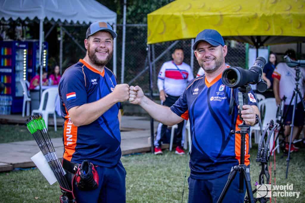 Twee boogschutters in bijpassende uniformen stoten hun vuist, staan naast handboogschietapparatuur en een richtkijker. Ze zijn buiten bij een sportevenement, met tenten en andere deelnemers op de achtergrond.