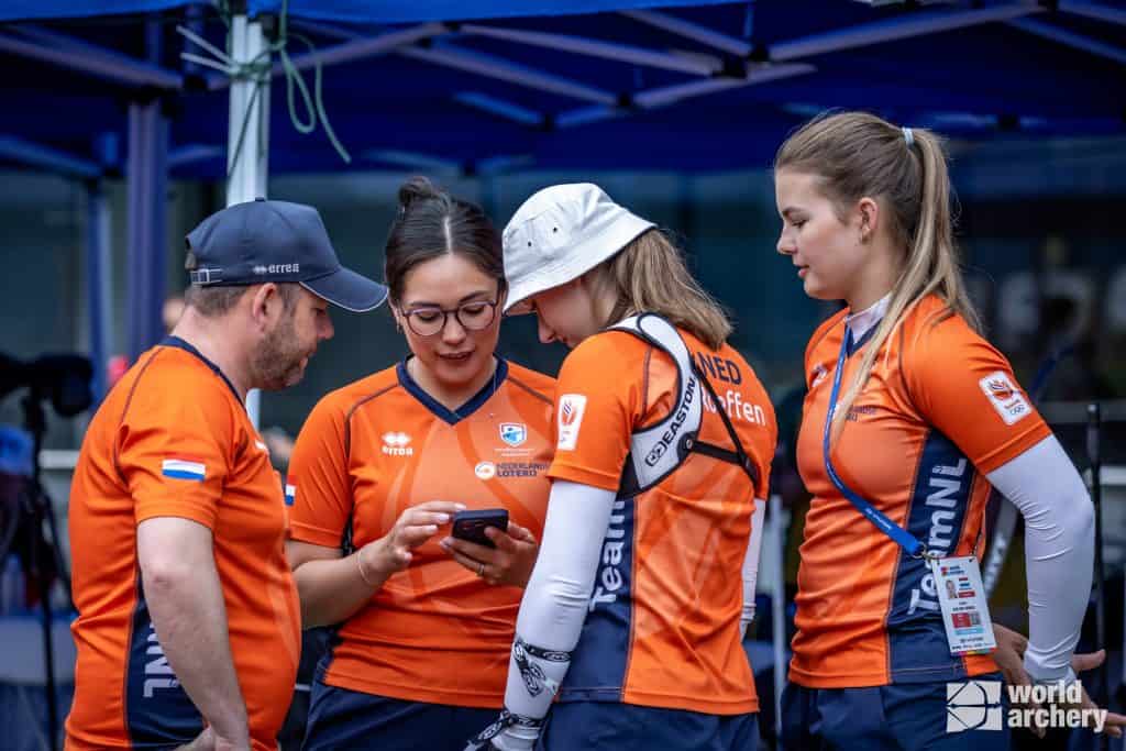 Vier boogschutters in oranje Team NL-uniformen staan samen onder een blauwe baldakijn, waarbij drie van hen gericht zijn op een smartphone die één van de boogschutters vasthoudt. Rechtsonder is een "World Archery" -logo zichtbaar, wat hun toewijding aan handboogsport benadrukt.