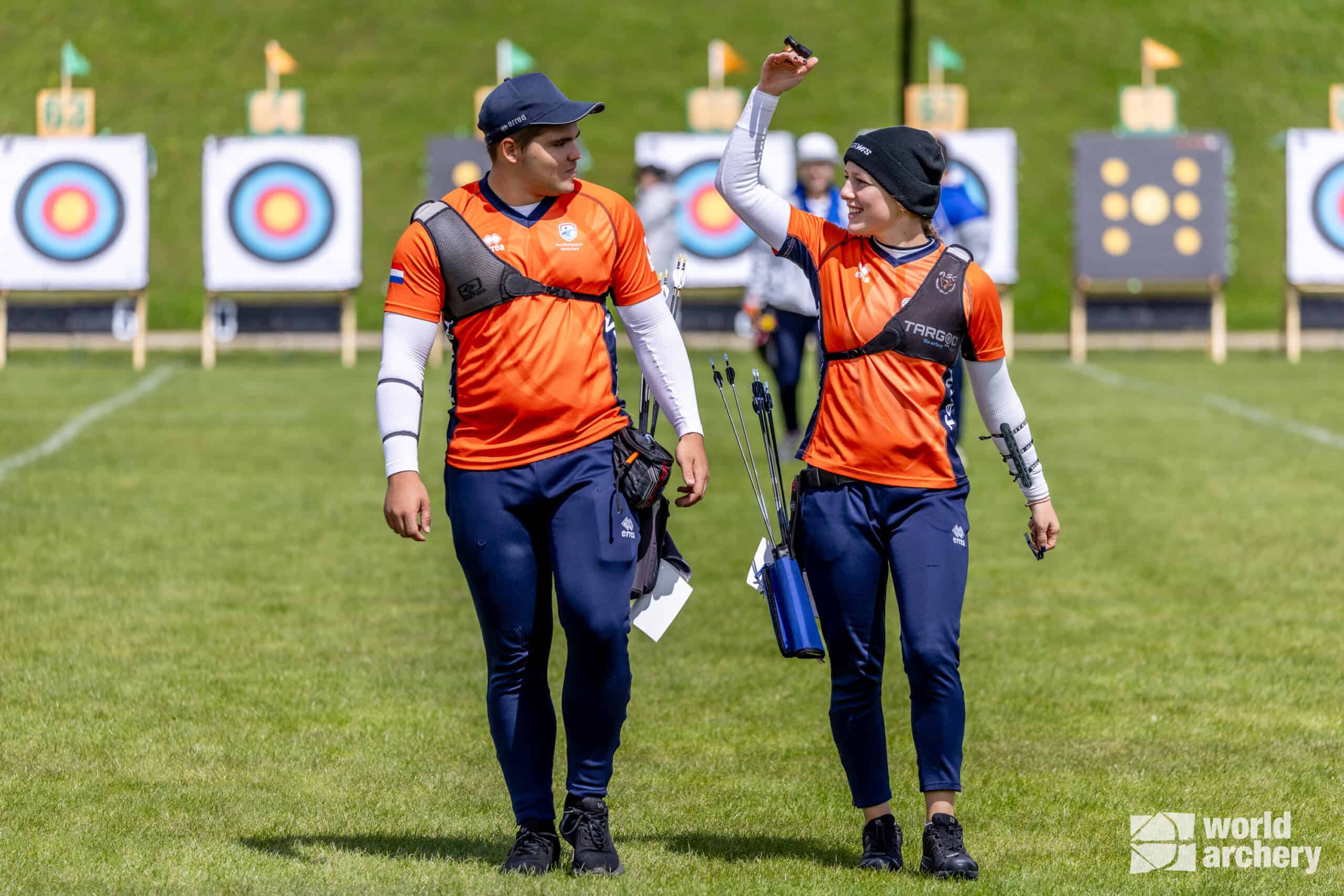 Twee boogschutters in oranje en marineblauwe kledij lopen op een grasveld met doelborden achter zich. Eén steekt een arm op en ze dragen allebei hun handboogsportuitrusting. Het World Archery-logo staat in de hoek.