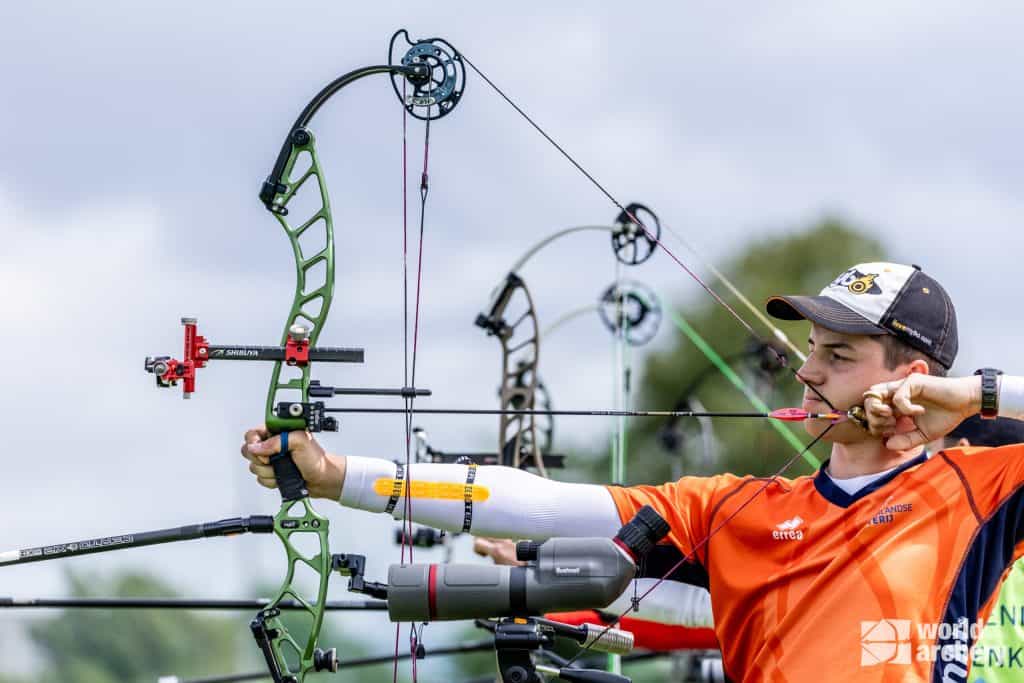 Een boogschutter in een oranje shirt richt een compoundboog tijdens een handboogsportwedstrijd in de open lucht. Op de achtergrond zijn andere boogschutters zichtbaar. De boogschutter draagt een vizierkap en er staat een telescoop op een statief vlakbij, klaar om de vlucht van elk pijl- en boogschot te volgen.