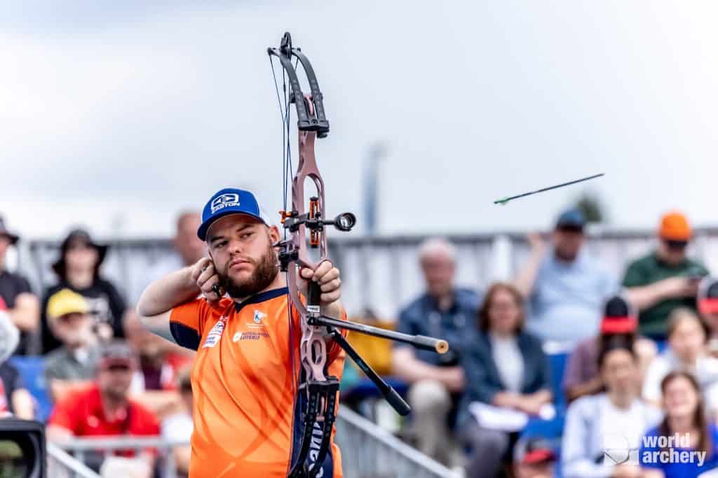 Een boogschutter, gekleed in een oranje shirt en een blauwe pet, richt een boog en schiet een pijl los tijdens een handboogsportwedstrijd, met een wazige menigte toeschouwers op de achtergrond.