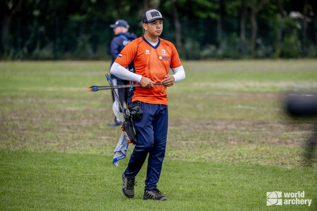 Een boogschutter in een oranje shirt en zwarte pet loopt over een grasveld met pijlen vast, bezig met handboogschieten, met een andere boogschutter op de achtergrond en het "world archery" -logo in de rechter benedenhoek.