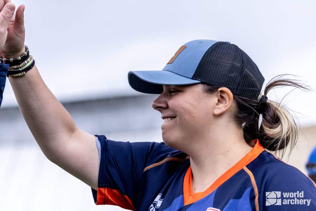 Een persoon die een marineblauw en oranje sporttrui en een pet draagt, lacht met opgeheven arm, alsof hij zojuist een succesvolle handboogschietsessie heeft afgerond. Het World Archery-logo is zichtbaar in de hoek van de afbeelding.