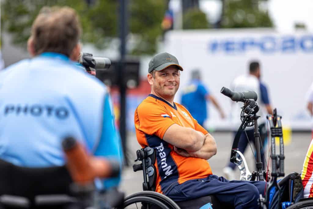 Een persoon die een oranje shirt en een pet draagt, glimlacht terwijl hij in een rolstoel zit, mogelijk opgewonden voor een handboogsport. Op de voorgrond is een andere persoon te zien in een lichtblauw shirt met "PHOTO" op de achterkant gedrukt.