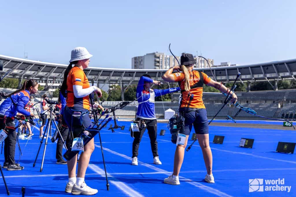 Boogschutters in uniform bereiden zich voor om pijlen te schieten met hun pijl en boog tijdens een wedstrijd op een blauw veld met een leeg stadion op de achtergrond. In de hoek is een officieel evenementlogo te zien.