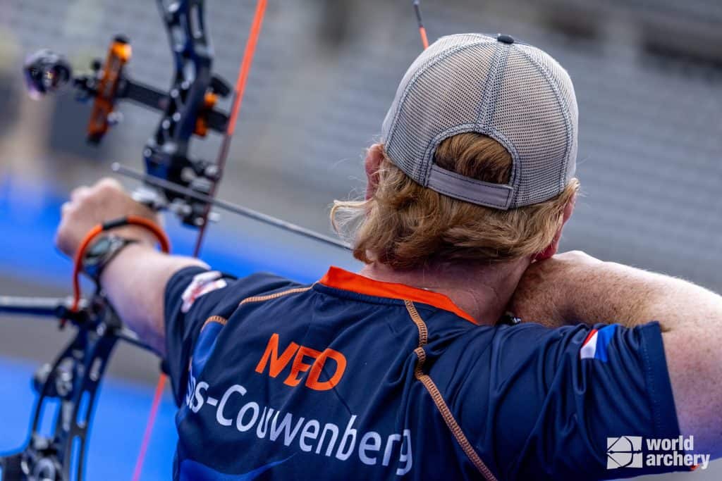 Een persoon met rood haar en een pet draagt, richt een pijl en boog tijdens een boogschietevenement. Op de achterkant van de blauwe trui staat "NED" en "Couwenberg." Rechtsonder is het World Archery-logo zichtbaar, wat de handboogsportcompetitie benadrukt.