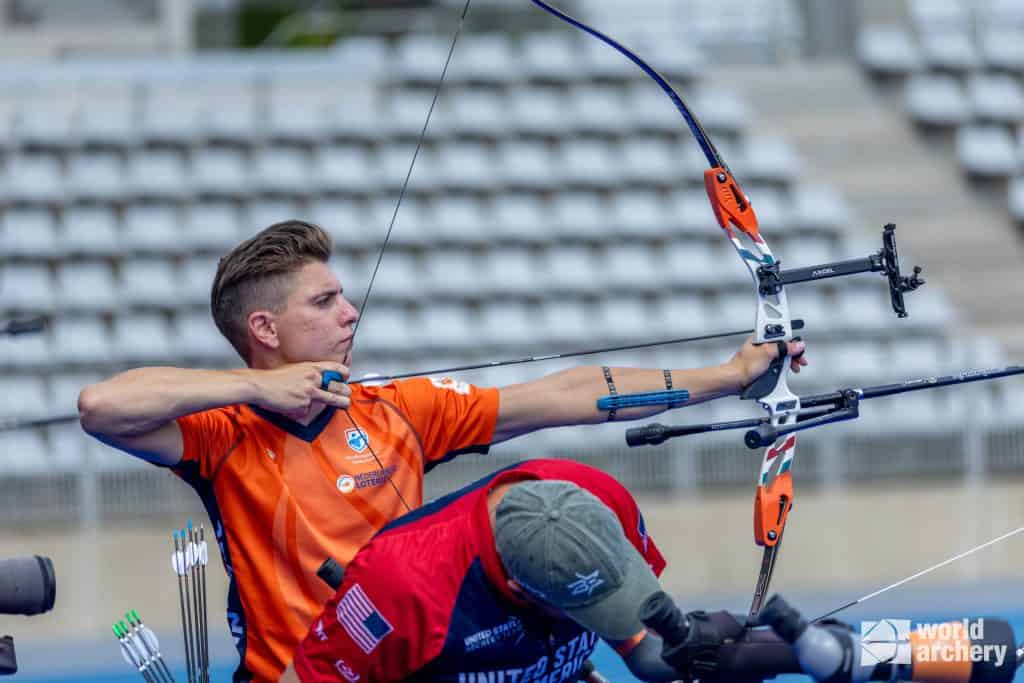 Een boogschutter in oranje shirt richt een pijl tijdens een handboogsportwedstrijd, terwijl een andere persoon op de voorgrond uitrusting lijkt aan te passen. Het evenement wordt georganiseerd door World Archery.