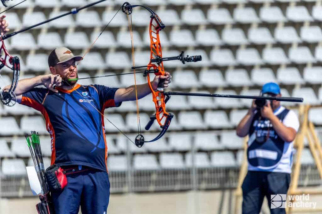 Een boogschutter die een compoundboog richt tijdens een handboogschietwedstrijd in een stadion, met een fotograaf die het moment op de achtergrond vastlegt.