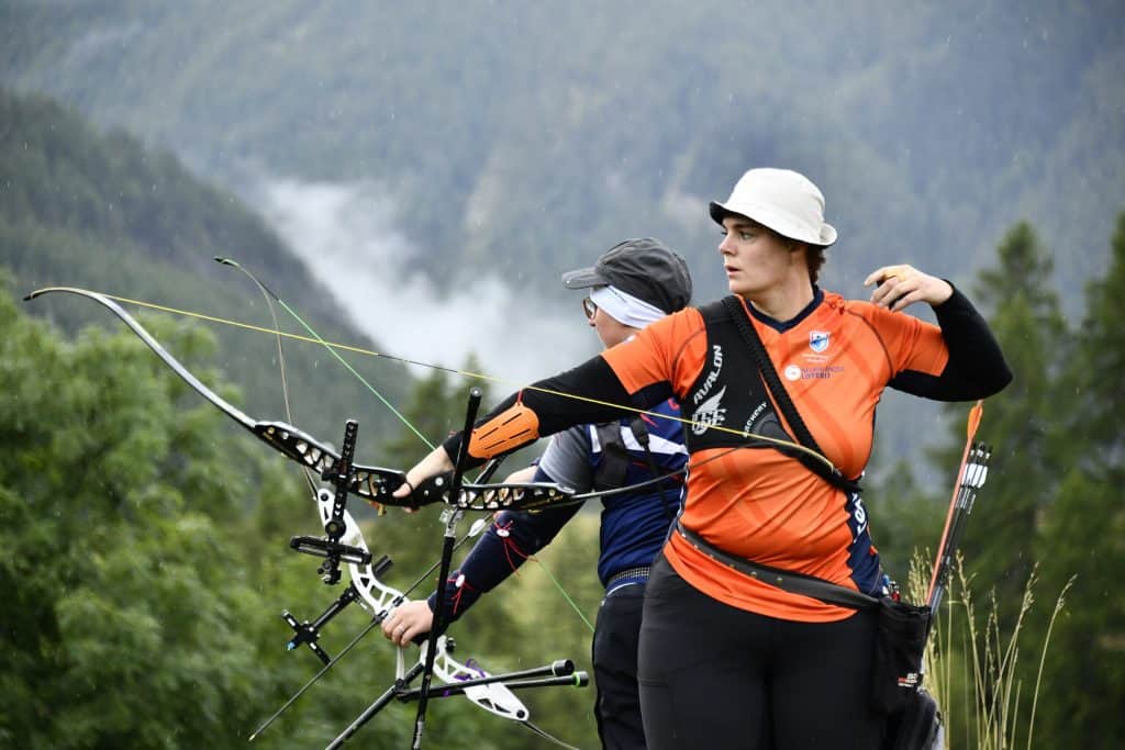 Twee boogschutters beoefenen handboogsport in een bergachtig gebied, de een draagt een oranje shirt en de ander in donkerblauw. Beiden gebruiken recurve boog omgeven door weelderig groen en met mist bedekte heuvels.
