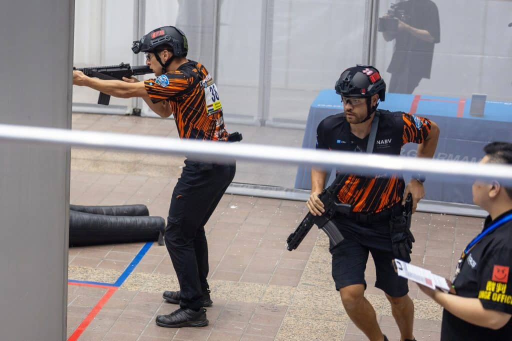 Twee personen in tactische uitrusting en helmen nemen deel aan een schietwedstrijd. De een richt een geweer vanuit dekking, terwijl de ander naar voren gaat. Op de voorgrond staat een waarnemer met een klembord, die doet denken aan handboogsport, maar dan met moderne vuurwapens in plaats van traditioneel handboogschietmateriaal.
