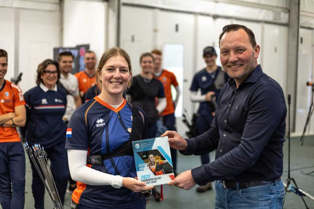 Een persoon in sportkleding ontvangt een onderscheidingsbordje van een ander in een casual trui, terwijl op de achtergrond een groep mensen toekijkt die mogelijk hun prestaties in de handboogsport vieren.