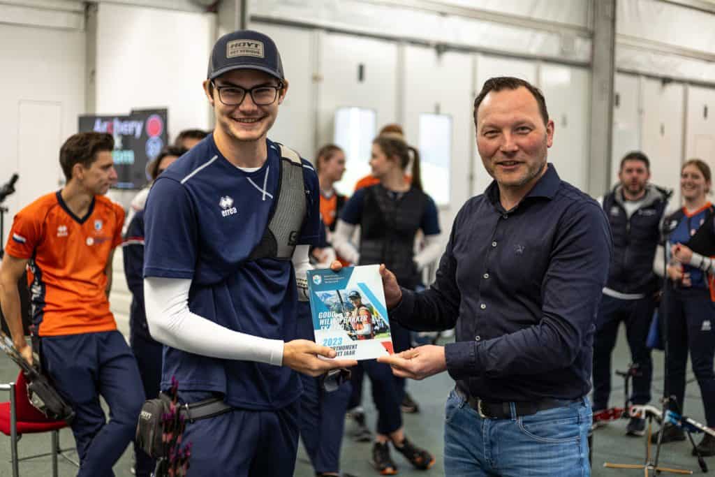 Twee mannen staan binnen, één in sportkleding met een pijl en een certificaat in de hand, de ander biedt het certificaat aan terwijl op de achtergrond mensen en handboogschietapparatuur verschijnen.