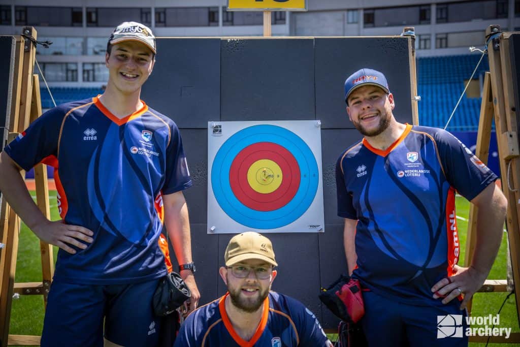 Drie boogschutters in teamuniformen staan rond een doelwit met pijlen op een boogschietbaan en glimlachen naar de camera. Het logo "world archery" is zichtbaar in de hoek van de afbeelding en toont hun passie voor handboogsport.