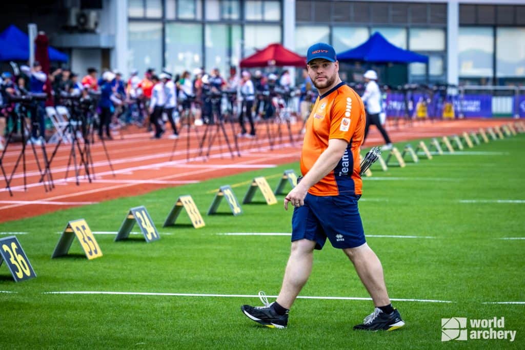 Een man in een oranje shirt en blauwe korte broek loopt op een atletiekveld met genummerde doelen. Hij houdt pijlen vast en heeft een boogschietuitrusting op zijn rug, waarmee hij duidelijk aan het handboogschieten doet. Het veld is ingericht voor een boogschietevenement.
