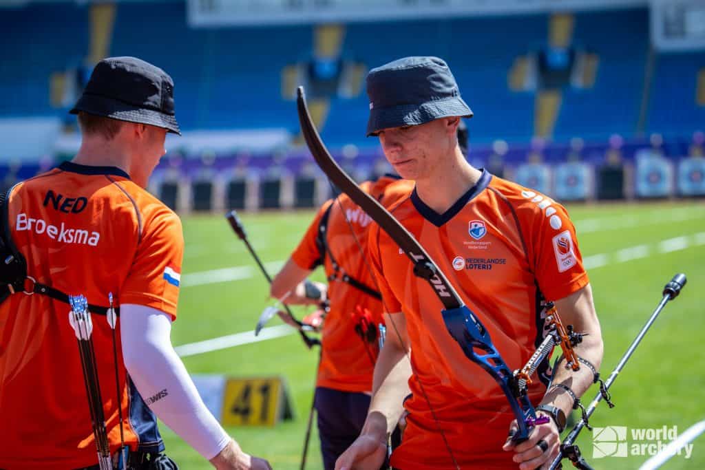 Op een veld met baannummer 41 zijn twee boogschutters in oranje shirts en hoeden, voorzien van pijl en boog, te zien. Ze bereiden zich voor op een handboogschieten in een stadionsetting.