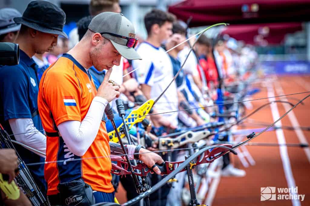 Boogschutters die in een rij staan, bereiden zich voor om te schieten tijdens de handboogsportcompetitie. De boogschutter op de voorgrond, gekleed in een oranje trui en pet, concentreert zich aandachtig met zijn hand voor zijn mond terwijl hij zijn boog vasthoudt.